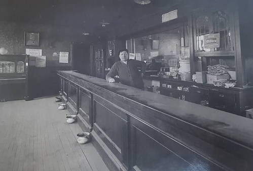 A historic black-and-white photo of a man standing behind a long bar in an old-fashioned establishment.