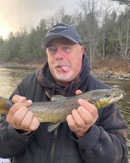 A man in a cap holds a trout, making a playful face by sticking out his tongue, with a river and trees in the background.