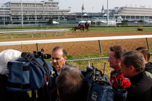 A group of reporters interviews a man at a horse racing track, with horses and a grandstand in the background.