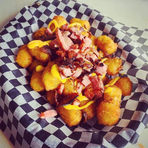 A serving of crispy tater tots topped with chopped bacon and drizzled with yellow mustard, served in a checkered paper basket.