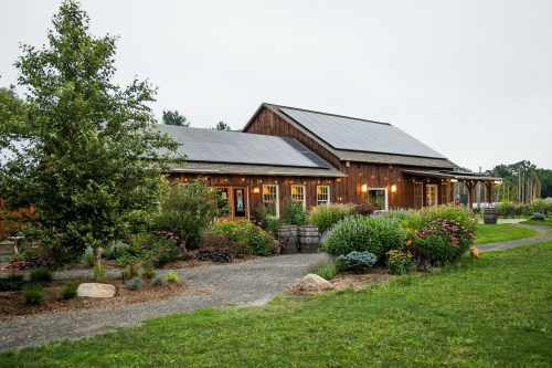 A rustic wooden building surrounded by lush gardens and a gravel path, under a cloudy sky.