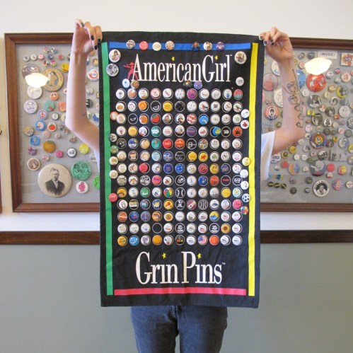 A person holds a black banner displaying colorful pins labeled "American Girl" and "Grin Pins," with a pin collection in the background.