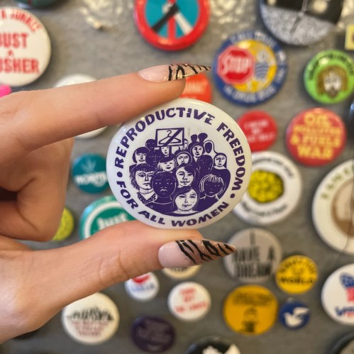 A hand holds a button that reads "Reproductive Freedom for All Women," surrounded by various other buttons.
