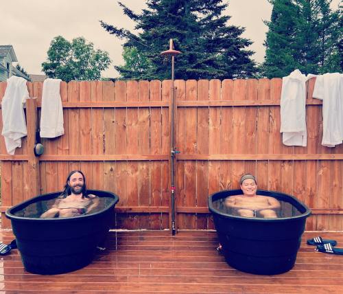 Two people relax in separate outdoor hot tubs on a wooden deck, surrounded by a wooden fence and trees.