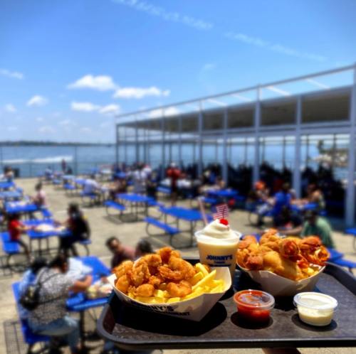 A tray with shrimp, fries, and dipping sauces in front of a waterfront dining area with people enjoying their meals.