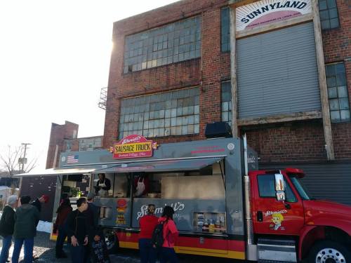 A sausage food truck parked outside a brick building, with people lined up to order food.