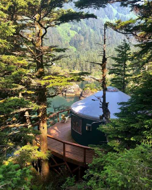 A cozy yurt nestled among tall trees, overlooking a serene body of water and lush green mountains.