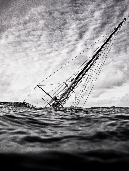 A partially submerged sailboat tilts in choppy waters under a cloudy sky, creating a dramatic black-and-white scene.