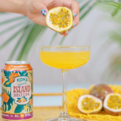 A cocktail glass filled with yellow liquid, garnished with a halved passion fruit, alongside a can of Kona Island Seltzer.