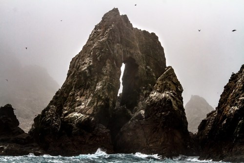 A rugged rock formation with a natural arch rises from turbulent ocean waters under a misty sky.