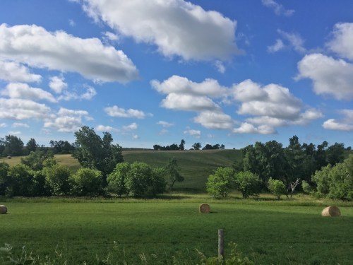 A scenic landscape featuring green fields, rolling hills, trees, and fluffy clouds in a blue sky.