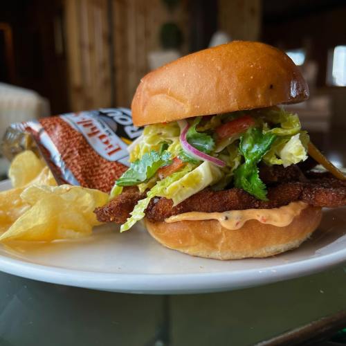 A crispy chicken sandwich with lettuce, onions, and sauce, served with potato chips on a white plate.