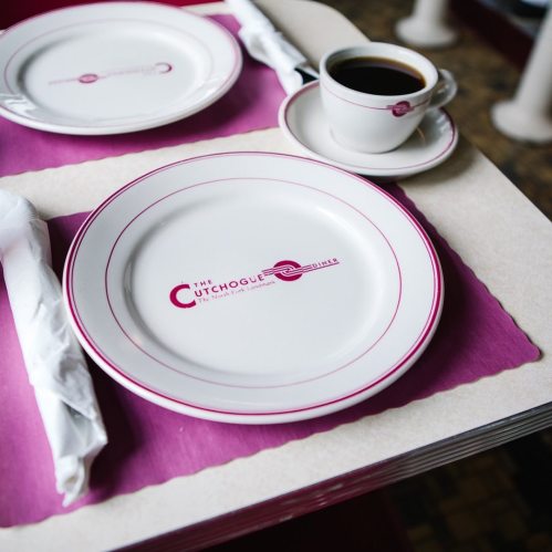 A table setting with a white plate, a coffee cup, and a purple placemat featuring the logo of "The Cutchogue."