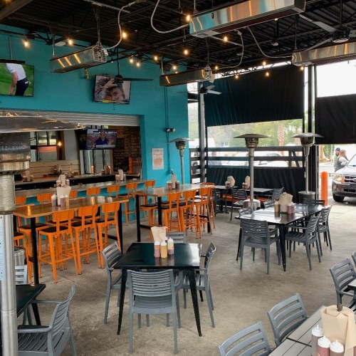 Outdoor dining area with colorful chairs, tables, heaters, and TVs, featuring a vibrant blue wall and a casual atmosphere.