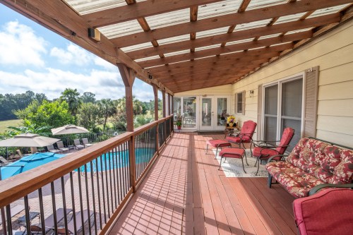 A spacious wooden deck with seating, overlooking a pool and lush greenery under a partly cloudy sky.