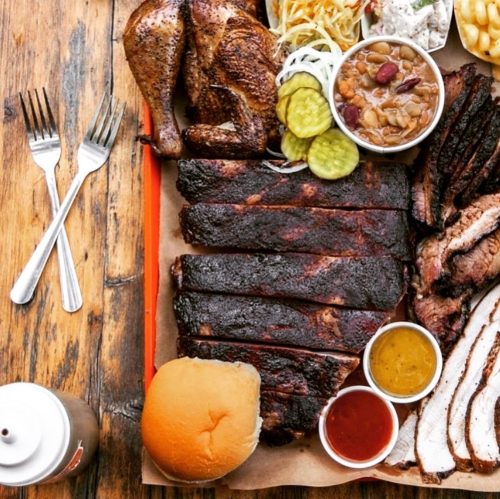 A platter of barbecue featuring ribs, chicken, coleslaw, pickles, beans, fries, and sauces, served with a bun.