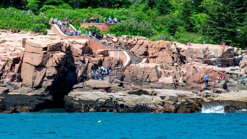 A rocky coastline with people walking on a path and stairs, surrounded by greenery and blue water.