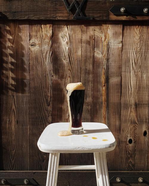 A glass of dark beer with foam spilling over, placed on a white stool against a rustic wooden background.