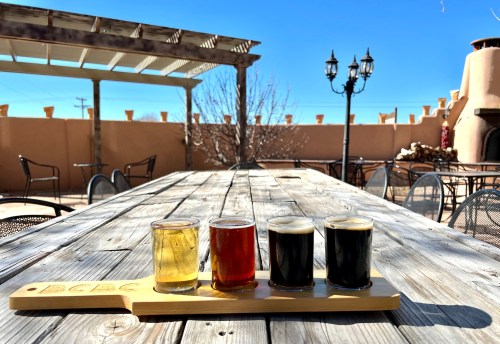 A wooden flight board with four beer samples sits on a rustic table outdoors, under a clear blue sky.