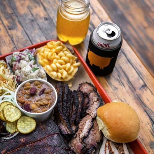 A BBQ platter with brisket, macaroni, coleslaw, beans, pickles, a bun, and a beer in a can on a wooden table.