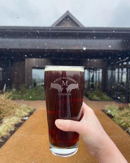 A hand holds a dark beer in a glass, with a rustic building and snowflakes in the background.