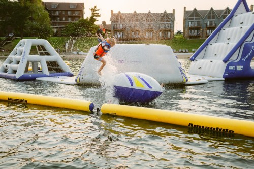A child jumps off a large inflatable water structure, splashing into the lake on a sunny day.