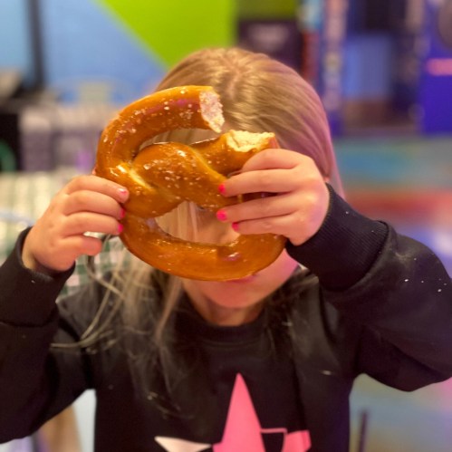 A child holds a large pretzel, taking a bite, with a colorful background.