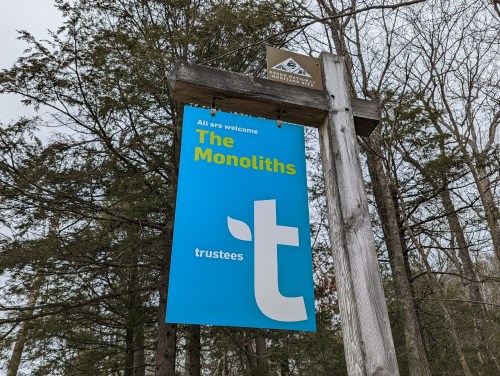 Signpost in a wooded area displaying "The Monoliths" and "trustees" in blue and white text.