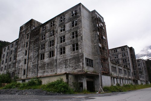 Abandoned concrete buildings with broken windows, surrounded by greenery and mountains under a cloudy sky.