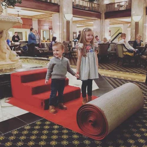 Two children stand on a red carpet in a hotel lobby, with a fountain and guests in the background.