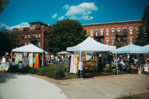A vibrant outdoor market with tents displaying clothing, surrounded by people and historic buildings under a blue sky.