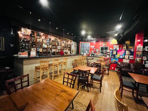 Cozy bar interior with wooden tables, chairs, and a well-stocked counter, featuring warm lighting and colorful decor.