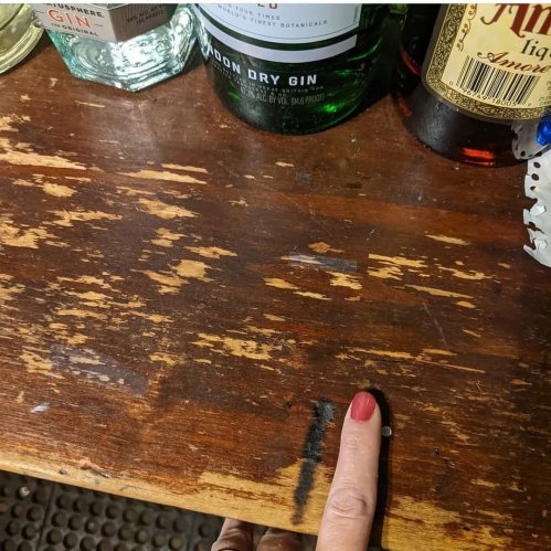 A hand with red nail polish points at a wooden surface with several bottles of gin and liquor in the background.