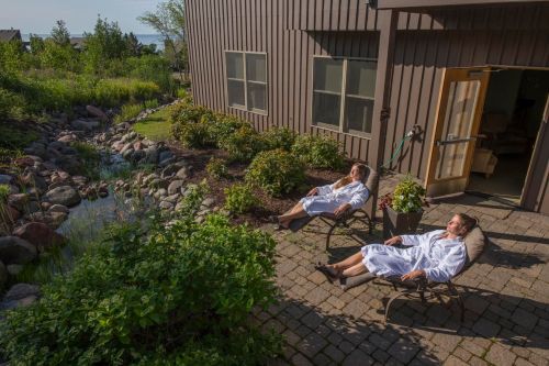 Two people in white robes relax on lounge chairs by a serene garden with a small stream and lush greenery.
