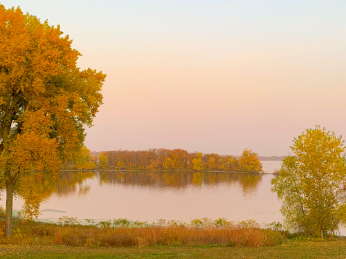A serene lakeside view at sunset, featuring vibrant autumn trees and calm water reflecting soft pastel colors.