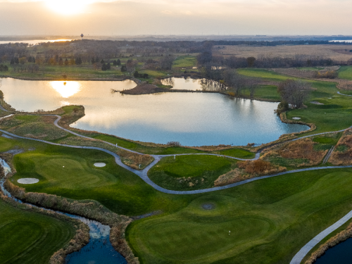 Aerial view of a golf course with a lake, lush green fairways, and a sunset in the background.