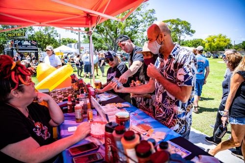 A group of people at a food stall, sampling sauces and enjoying a sunny outdoor event with tents and vendors in the background.