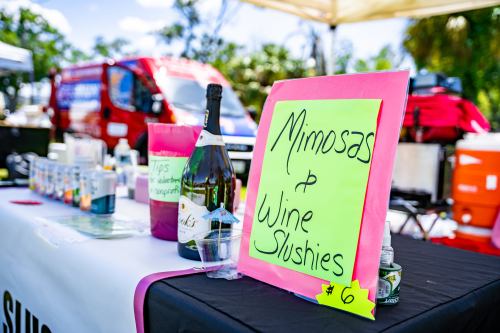 A colorful sign displays "Mimosas & Wine Slushies" for $6 at a vibrant outdoor market stall.