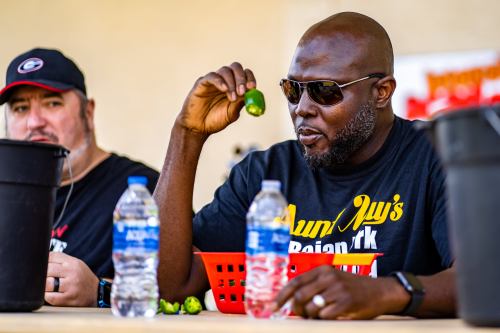 A man in sunglasses holds a pepper, sitting at a table with water bottles and another person in the background.