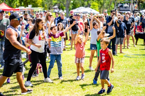 A lively crowd of children and adults enjoying an outdoor event, dancing and celebrating in a sunny park.