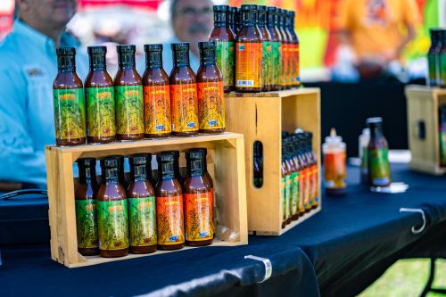 Bottles of sauce displayed on a table, with wooden crates and colorful labels in a market setting.
