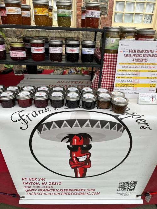 Display of jars filled with salsa, pickled vegetables, and preserves at a market stall, featuring a cartoon chili pepper logo.