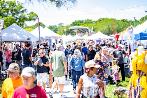 A bustling outdoor market with crowds walking between vendor tents on a sunny day.