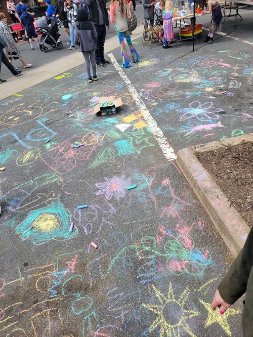 Colorful chalk drawings cover a pavement, with people and children playing in a lively outdoor setting.