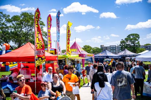A lively outdoor festival scene with colorful tents, food stalls, and crowds enjoying the sunny day.