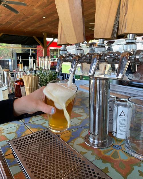 A hand pours a foamy beer into a glass at a bar with taps and colorful tile.