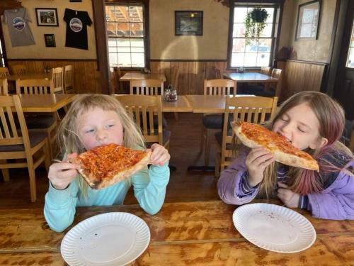 Two children sitting at a wooden table in a restaurant, happily eating large slices of pizza.