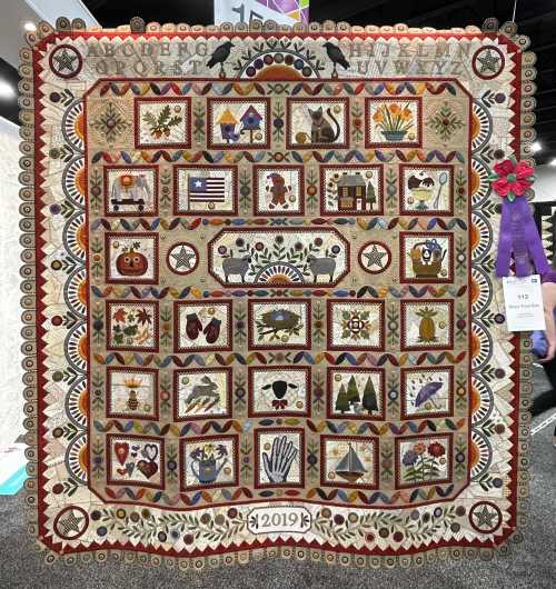 A colorful quilt featuring various embroidered blocks with patterns of animals, houses, and nature, framed with decorative borders.