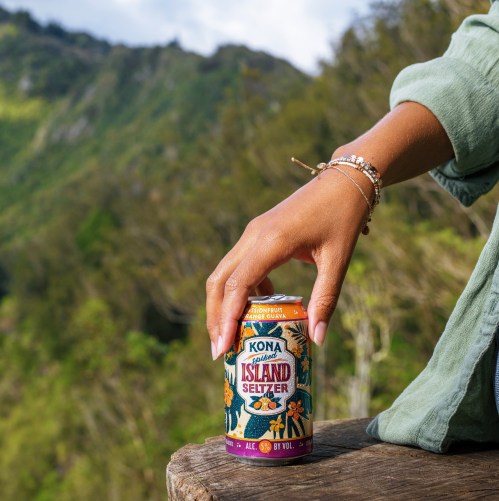 A hand holding a can of Kona Island Seltzer, set against a scenic mountain backdrop.