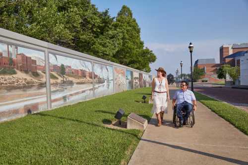 A woman in a hat walks beside a man in a wheelchair along a mural-adorned path by a river.